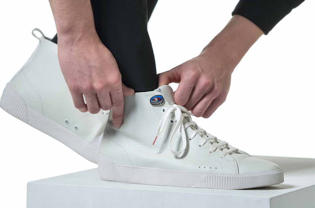 Modelo ajustando con sus manos unos zapatos blancos de media caña que llevan engastados la insignia con un diamante del Spotify Camp Nou sobre fondo blanco.