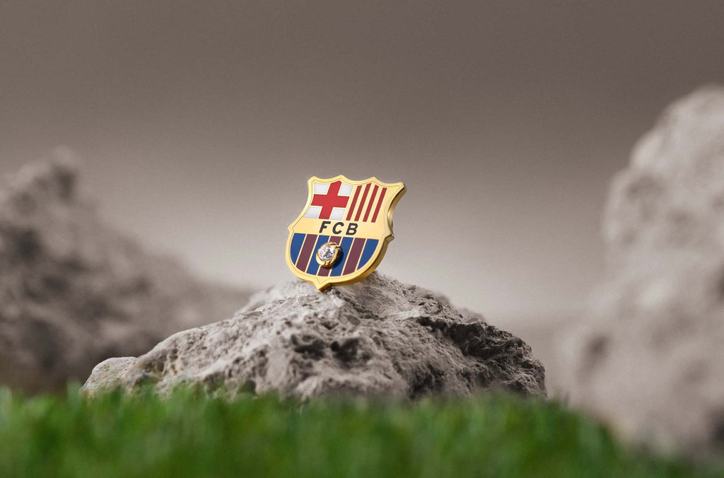 La emblemática insignia del escudo del FC Barcelona, presentada sobre una piedra en forma triangular rodeada de césped y otras piedras. La brillante insignia, bañada en oro y esmalte, resalta, reflejando la grandeza del club