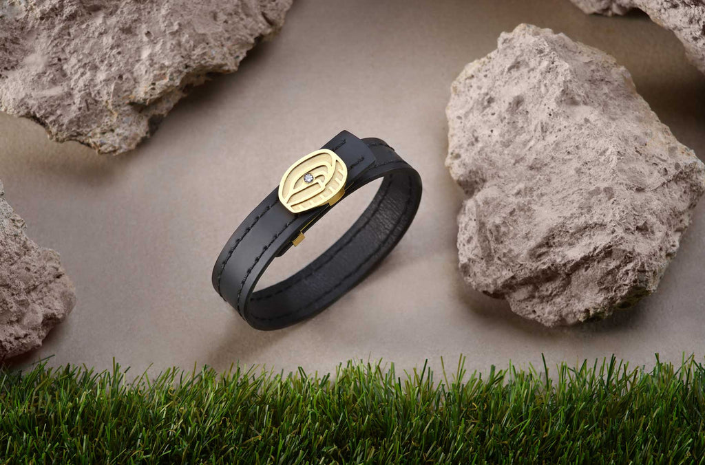 La pulsera de elegante diseño de cuero genuino de color negro y bañada en oro, un verdadero emblema del Spotify Camp Nou. Rodeada de césped y piedras que reflejan su esencia, con un deslumbrante diamante en su centro.