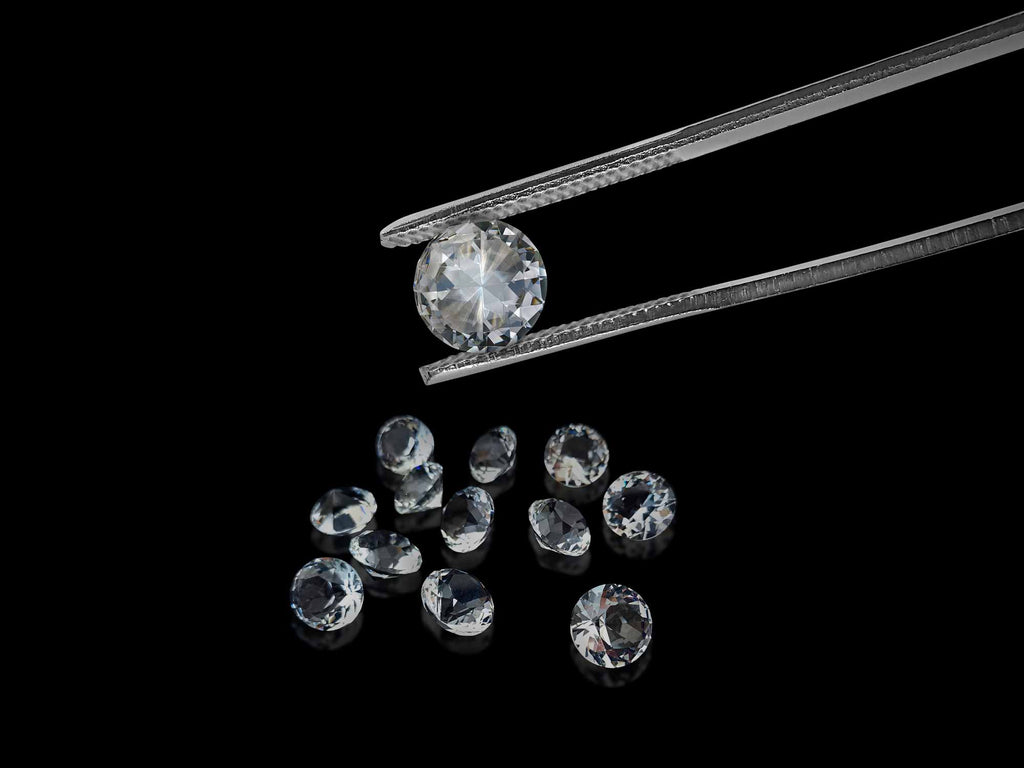 Pinzas sujetando un diamante en primer plano, con otros diamantes difuminados en un fondo negro.