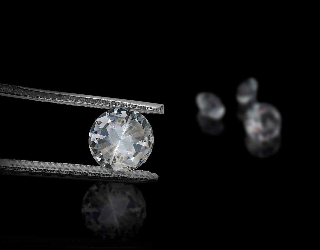 Pinzas de joyería sostienen un diamante, con otros diamantes en segundo plano difuminados sobre fondo negro.