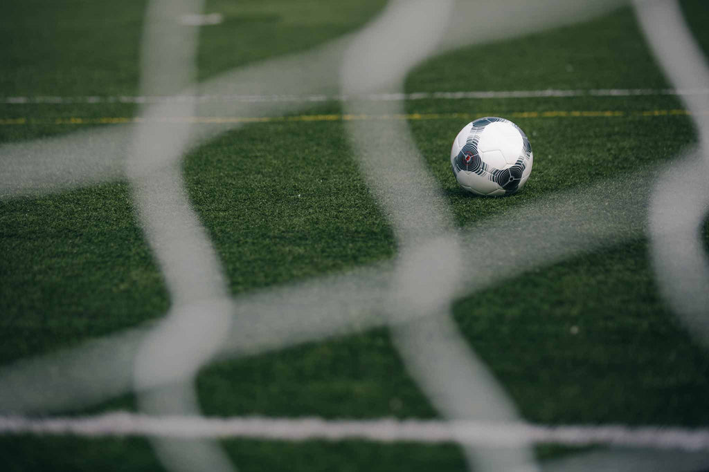 Pelota de fútbol en segundo plano, enfocada y nítida, con una red de fútbol desenfocada en el primer plano, vista desde detrás de la red.